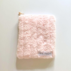 May include: A rectangular, pale pink faux fur pouch with a gold-coloured zip and a small white label reading "Sew Sunday". The pouch has a soft, fluffy texture, and is likely for storing small items.