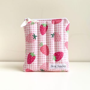 May include: A quilted pouch featuring a pink gingham check pattern and a strawberry print. The strawberries are a bright pink with green stems. The pouch has a white zip and a small white bow. The text "Sew Sunday" is visible on the bottom right.