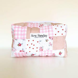 May include: A patchwork cosmetic bag with a variety of fabrics. The bag features pink gingham, floral, and heart patterns. The text "Sew Sunday" is printed on a white label. The bag has a white zip.