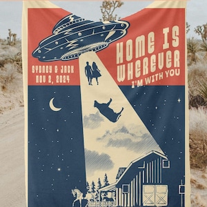 May include: A personalized blanket with a retro design featuring a UFO, a couple, and a cow being beamed up. The blanket has the text "Home is Wherever I'm With You" and "Sydney & Jack Nov 2, 2024". The blanket is cream-colored with a desert background.