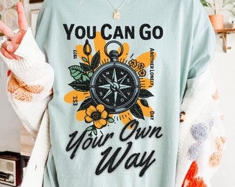 May include: A light blue t-shirt with a compass design and the text "You Can Go Your Own Way" in black. The compass is surrounded by flowers and leaves in yellow, green, and orange. The text "Another Lonely Day" is written vertically on the right side of the compass.