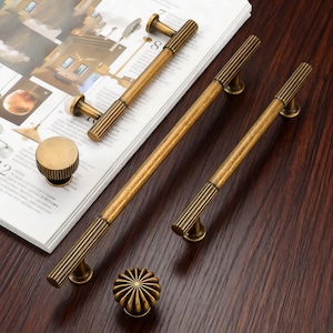 May include: A selection of antique brass cabinet hardware, including handles and knobs. The handles have a ribbed design and come in various sizes. The knobs feature a round, fluted design. The hardware is displayed on a wooden surface and a magazine.
