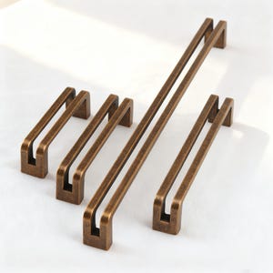 Könnte beinhalten: Eine Auswahl an bronzefarbenen Schrankgriffen in verschiedenen Größen. Die Griffe haben ein rechteckiges Design mit einem Doppelstangengriff. Die Oberfläche hat ein leicht gealtertes Aussehen, das einen Vintage-Stil suggeriert. Maße in cm.