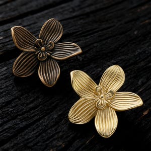 May include: Two decorative flower-shaped cabinet knobs. One is a bronze color, and the other is gold. Each knob has five petals with detailed linear grooves and a small center. The knobs are on a dark wood surface.
