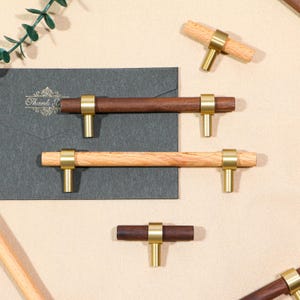 May include: Wooden cabinet pulls with brass accents. The pulls feature a T-bar design with a cylindrical wooden handle and a brass ring and base. One pull is dark brown, and the other is light brown.