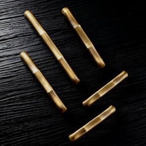 May include: Five brass cabinet pulls with a brushed finish are arranged diagonally on a dark wood surface. Each pull has a cylindrical shape with a contrasting band in the center.