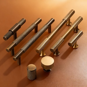 May include: A collection of antique brass cabinet hardware, including various sizes of bar pulls and knobs. The pulls feature textured and grooved designs, with a warm, aged finish. The hardware is arranged on a brown surface.