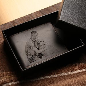 May include: A dark brown leather wallet with a custom engraved image of a person and a dog inside a black gift box. The wallet is open, revealing the detailed engraving. The box is on a wooden surface.
