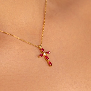 May include: A gold necklace with a cross pendant. The cross is adorned with red gemstones and a single white gemstone at the center. The necklace is displayed on a person's skin.