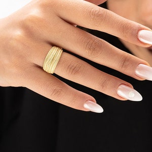 May include: A gold band ring with a textured design. The ring is worn on the index finger of a person's hand.