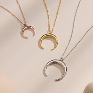 May include: Three crescent moon necklaces in rose gold, gold, and silver. Each pendant hangs from a delicate chain. The pendants are smooth and curved, with a polished finish. The necklaces are displayed on a light-colored surface.