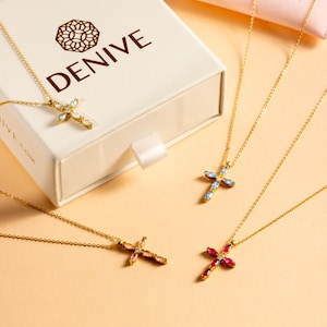 May include: Four gold necklaces with cross pendants featuring different colored gemstones. The necklaces are displayed on a light beige surface. The cross pendants are adorned with blue, pink, and red gemstones.
