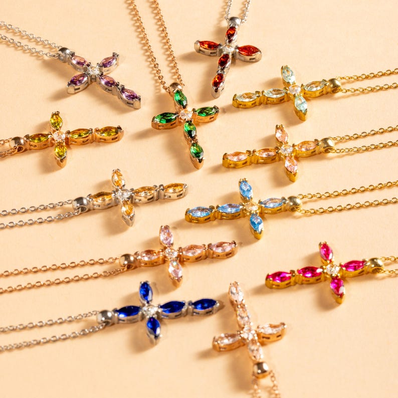 May include: A collection of gold cross necklaces with different colored gemstones. The gemstones are in a marquise shape and are set in a pave setting. The necklaces are all on a white background.