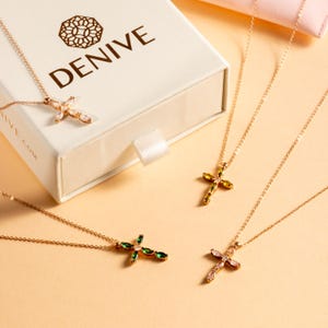 May include: Four gold necklaces with cross pendants featuring different colored gemstones. The crosses are adorned with clear and colored marquise-shaped stones. The necklaces are displayed on a light beige surface.