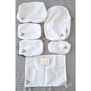 Könnte beinhalten: Ein Set weißer Reise-Organizer, darunter ein Rucksack, mehrere Reißverschlusstaschen und ein Kordelzugbeutel. Jedes Teil hat ein schwarzes Logo in der Ecke. Die Artikel sind auf einer grauen Oberfläche angeordnet.