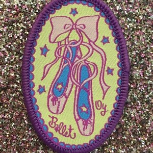 Op de afbeelding: Een geborduurde patch in paars en geel met een paar roze balletten met een strik erop. De patch heeft een witte achtergrond met sterren en het woord "Ballet" in paars.