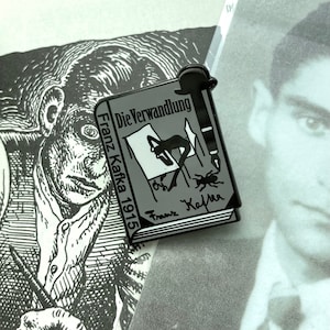 May include: A gray enamel pin shaped like a book with the title "Die Verwandlung" by Franz Kafka, 1915. The pin features a black and white illustration of a man drawing a beetle.
