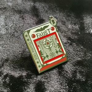 May include: A silver and red enamel pin shaped like a book with the title "FAUST Goethe 1808" and a stylized image of an eye inside a pentagram.