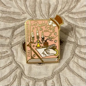 Puede incluir: Un pin de esmalte rosa con una portada de libro con el título "Daddy Long Legs" de Jean Webster, publicado en 1912. El pin representa una escena de una mano escribiendo una carta con una pluma estilográfica, un candelabro y un tintero.