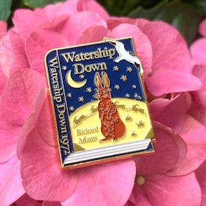 May include: A gold-coloured enamel pin featuring the cover of the book Watership Down by Richard Adams. The pin depicts a rabbit standing on a hill under a crescent moon and stars. The book title and author's name are visible on the pin.