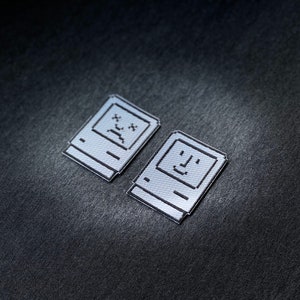 May include: Two silver-coloured iron-on patches depicting a retro computer with a happy face and a sad face. The patches are on a dark grey background.
