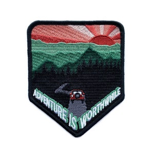 May include: Black and green embroidered patch featuring a sunset over mountains and a jeep driving on a winding road. The text "Adventure is worthwhile" is written in white on a green background.
