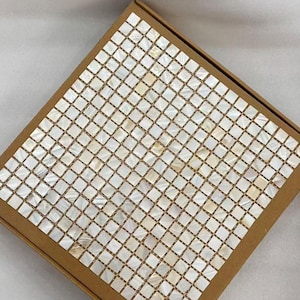 Puede incluir: Una caja cuadrada marrón con un diseño de mosaico. Las baldosas son pequeñas, cuadradas y blancas con un brillo nacarado. Las baldosas están dispuestas en un patrón de cuadrícula, creando una superficie texturizada. La caja está sobre un fondo blanco.