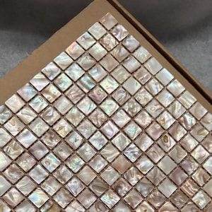 Op de afbeelding: Een mozaïektegelblad bestaande uit kleine, vierkante tegels. De tegels hebben een parelmoerachtige, iriserende kwaliteit met tinten wit, beige en bruin. De tegels zijn gerangschikt in een rasterpatroon, waardoor een glinsterend oppervlak ontstaat.