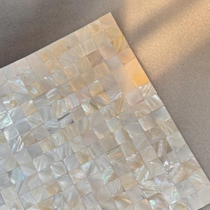 Puede incluir: Azulejo de mosaico cuadrado compuesto por numerosos cuadrados pequeños, iridiscentes y blanquecinos. El azulejo tiene un brillo nacarado, que refleja la luz con toques de amarillo y azul. El mosaico está sobre un fondo neutro.