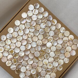 Puede incluir: Un cuadrado de baldosas de mosaico redondas e iridiscentes en una caja de cartón. Las baldosas son una mezcla de blanco, crema y beige, creando un efecto nacarado. La caja es de color marrón natural y las baldosas están dispuestas en un patrón de cuadrícula.