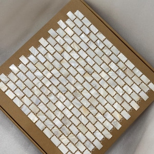 Puede incluir: Una caja de baldosas de mosaico rectangulares e iridiscentes. Las baldosas están dispuestas en un patrón de ladrillo, con un brillo nacarado. Las baldosas están sobre una base de cartón marrón.