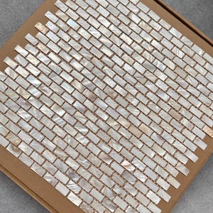 Può includere: Un foglio di piastrelle a mosaico rettangolari con finitura madreperla. Le piastrelle sono disposte a motivo a spina di pesce e sono inserite in una cornice di cartone. Le piastrelle iridescenti riflettono la luce, creando un effetto scintillante. Ideale per progetti di home decor.