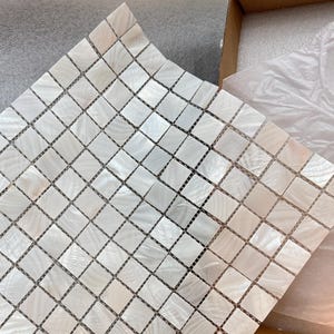 Könnte beinhalten: Ein quadratisches Blatt Mosaikfliesen mit Perlmutt-Finish. Die kleinen, quadratischen Fliesen sind in einem Rastermuster angeordnet. Die Fliesen sind eine Mischung aus Weiß und Off-White, mit einem subtilen Schimmer. Das Blatt ist teilweise aus einem Karton.