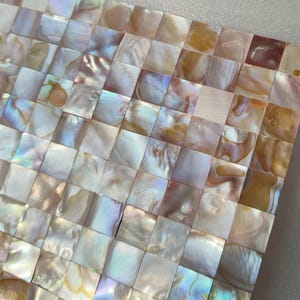 May include: Close-up of a square mosaic tile made of small, iridescent, square pieces. The tiles have a pearlescent sheen with colours ranging from white and gold to hints of blue and pink. The mosaic is set against a neutral background.
