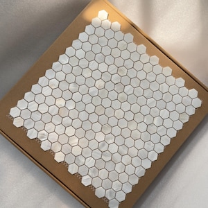 May include: A box of white, hexagonal mosaic tiles. The tiles have a pearlescent sheen and are arranged in a grid pattern. The box is made of brown cardboard and is set against a white background.