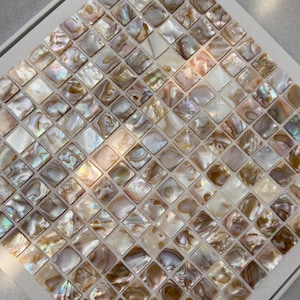May include: A square mosaic tile sheet featuring numerous small, individual square pieces of iridescent mother-of-pearl. Each piece displays a unique, natural pattern with varying shades of white, cream, beige, and soft brown with a pearlescent finish.