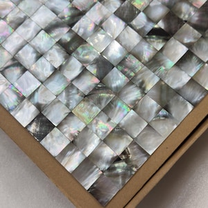 Peut inclure: Gros plan sur une mosaïque de carreaux carrés décoratifs. La mosaïque est composée de petits carreaux irisés en nacre disposés en motif tissé. Les carreaux reflètent la lumière avec une gamme de couleurs, dont le blanc, le gris et des reflets arc-en-ciel.