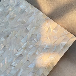 Pode incluir: Close-up de uma folha de azulejo de mosaico retangular. Os azulejos são retangulares, dispostos em um padrão de tijolo e feitos de material branco iridescente. A superfície reflete a luz, criando um efeito brilhante. O fundo é uma superfície neutra e texturizada.