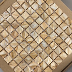 May include: A square sheet of mosaic tiles with a grid pattern. The tiles are a mix of beige and cream colors, with a pearlescent sheen. The tiles are set in a cardboard box.