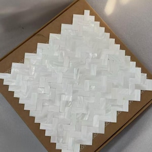 May include: A square mosaic tile sheet featuring a white herringbone pattern made of iridescent mother of pearl pieces. The individual rectangular tiles are arranged in a repeating woven design, creating a shimmering surface texture on a brown backing.
