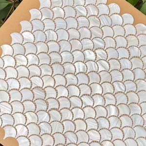 Op de afbeelding: Witte, waaiervormige mozaïektegels in een herhalend patroon. De tegels hebben een parelmoerglans en zijn geplaatst op een bruine achtergrond. Het ontwerp lijkt op visschubben, geschikt voor interieurprojecten.