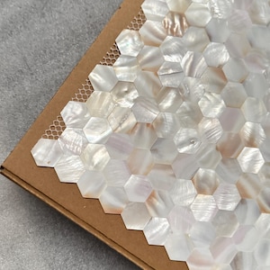 Puede incluir: Muestra de baldosas hexagonales de nácar. Las baldosas iridiscentes blancas y crema están dispuestas en un patrón de panal. Las baldosas están colocadas en una bandeja de cartón marrón con un respaldo de malla.