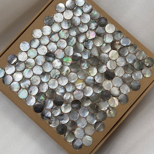 Puede incluir: Un cuadrado de baldosas redondas e iridiscentes de nácar colocadas en una caja de cartón. Las baldosas tienen una calidad brillante y nacarada con tonos de gris, plata y toques de otros colores. Las baldosas están dispuestas en un patrón de mosaico.
