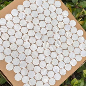 Puede incluir: Una lámina de azulejos de mosaico blancos y redondos dispuestos en un patrón de cuadrícula. Los azulejos tienen un brillo nacarado y están colocados sobre un respaldo marrón. El fondo presenta follaje verde.