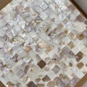 Puede incluir: Una lámina de mosaico cuadrado con un patrón tejido. Las baldosas son una mezcla de piezas de concha blanca, crema e iridiscente, creando un efecto brillante. El mosaico está dentro de un marco de cartón marrón.