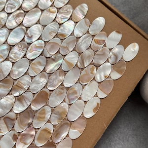 Puede incluir: Primer plano de una hoja de mosaico con numerosas baldosas ovaladas. Las baldosas tienen un acabado nacarado en tonos blancos, crema y beige. Las baldosas están dispuestas en un patrón de cuadrícula.