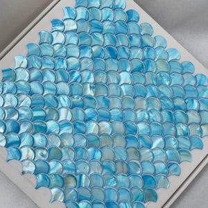 May include: A square of iridescent blue mosaic tiles arranged in a fish scale pattern. The tiles are a mix of light and dark blue, creating a shimmering effect. The tiles are set on a white backing.