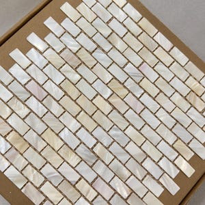Puede incluir: Una caja de baldosas de mosaico rectangulares en un patrón diagonal. Las baldosas son de color blanquecino con reflejos iridiscentes, sobre un respaldo de cartón marrón. Las baldosas miden aproximadamente 2,5 cm por 1,25 cm.