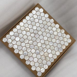 Puede incluir: Una caja de baldosas de mosaico hexagonales. Las baldosas son blancas con un brillo nacarado, dispuestas en un patrón de panal. Las baldosas están colocadas dentro de una caja de cartón marrón.