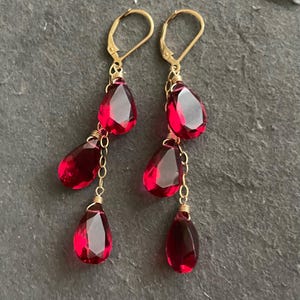 May include: Gold dangle earrings with three red teardrop-shaped crystals on each earring.